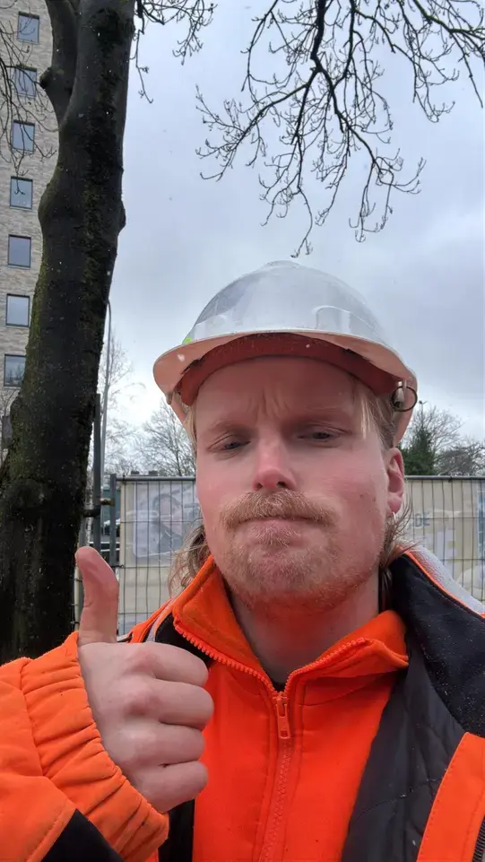 Selfie van een medewerker met witte helm en oranje werkjas die zijn duim opsteekt