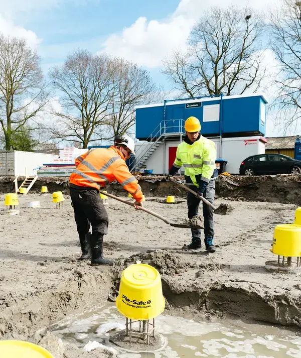 Twee medewerkers aan het werk op een bouwterrein met bouwketen op de achtergrond