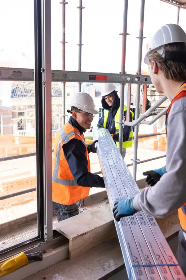 Medewerkers met helmen tillen een raamkozijn bij een steiger