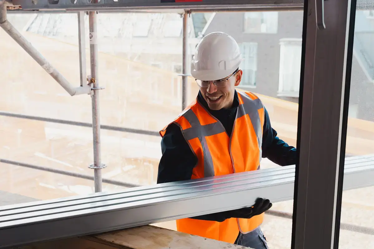 Medewerker met helm en veiligheidshesje tilt een raamkozijn op de bouwplaats